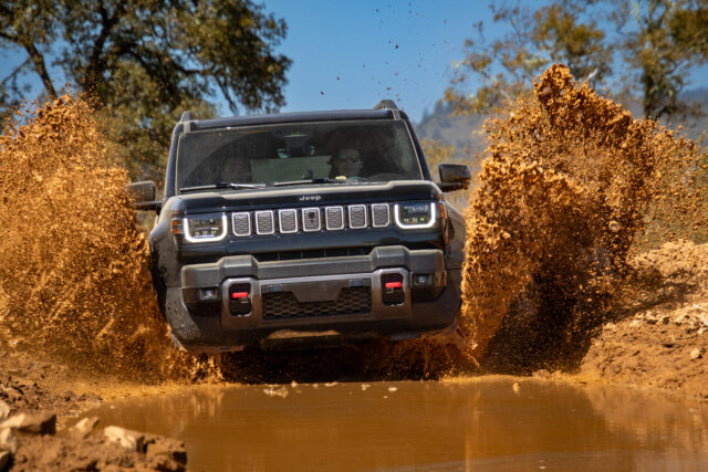 A black Jeep Recon SUV drives through a large muddy puddle, splashing water and mud high into the air, with trees and blue sky in the background.