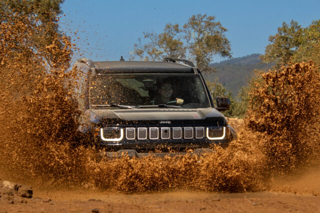 A black Jeep Recon drives through a large mud puddle, splashing muddy water high on both sides. Trees and hills are visible in the background under a clear blue sky.