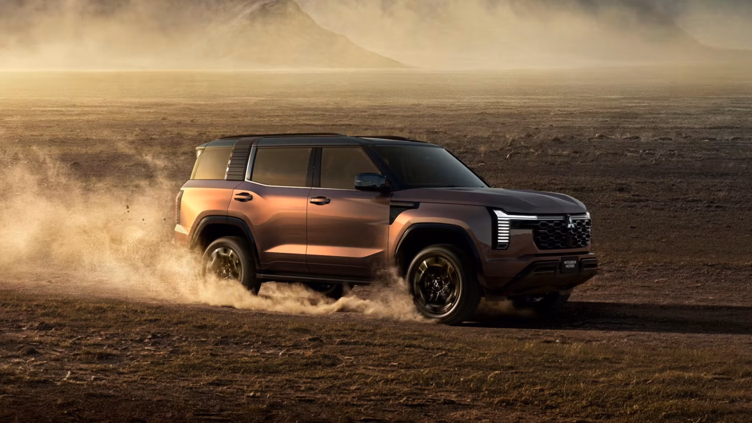 A rugged brown Mitsubishi Pajero drives through a dusty landscape, kicking up clouds of sand, with mountains and a hazy sky in the background.