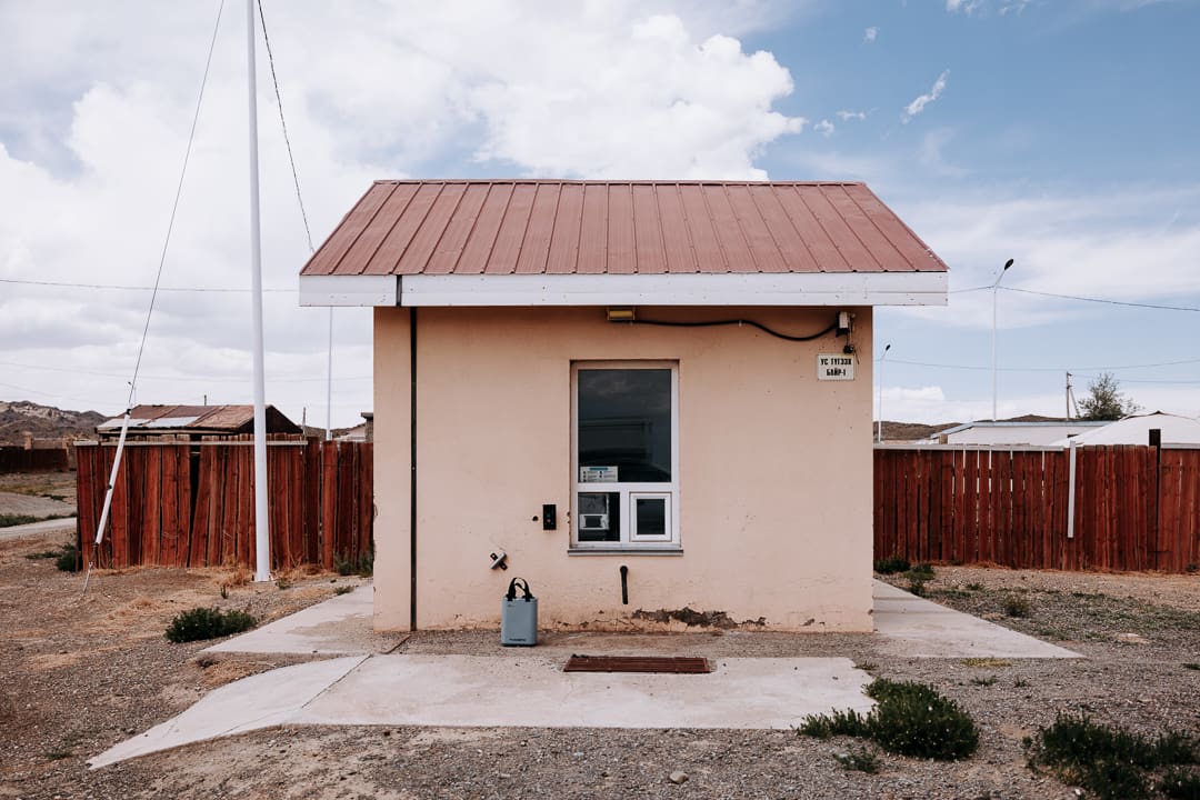 A small beige building with a red roof stands in front of a red wooden fence beneath a cloudy sky—an inviting scene reminiscent of overlanding Mongolia. A window and utility meter are visible on the front wall, while sparse vegetation surrounds the concrete entryway.