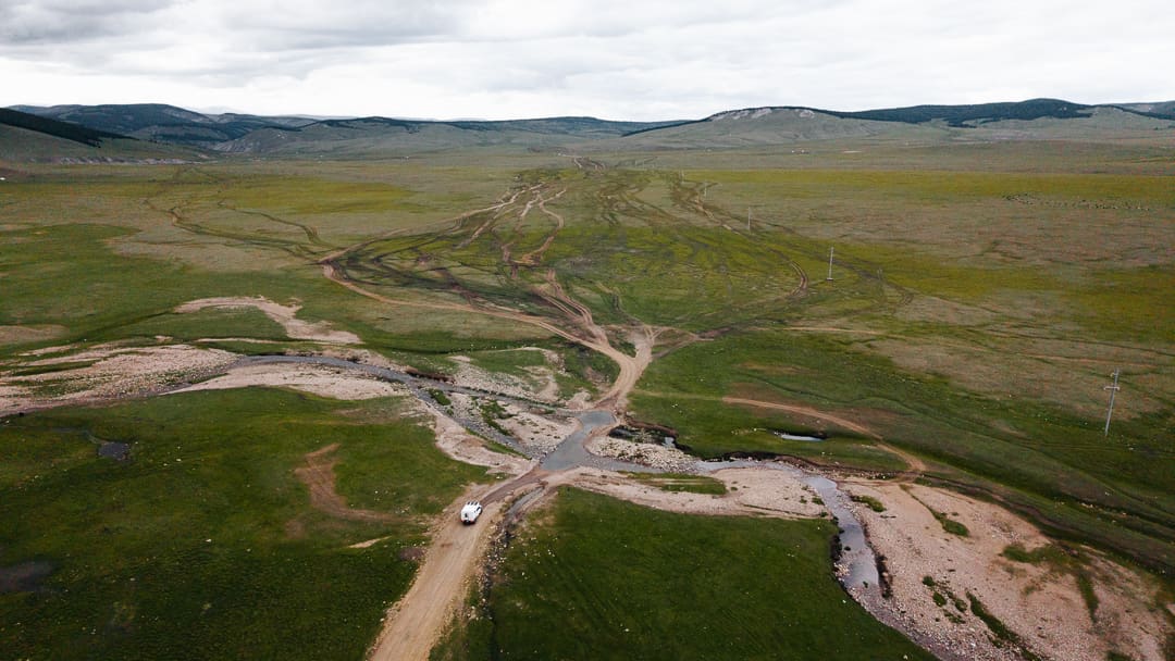 A white vehicle drives on a dirt road across a grassy plain with multiple dirt tracks and a small stream, surrounded by distant hills under a cloudy sky—capturing the spirit of overlanding Mongolia’s vast and rugged landscapes.