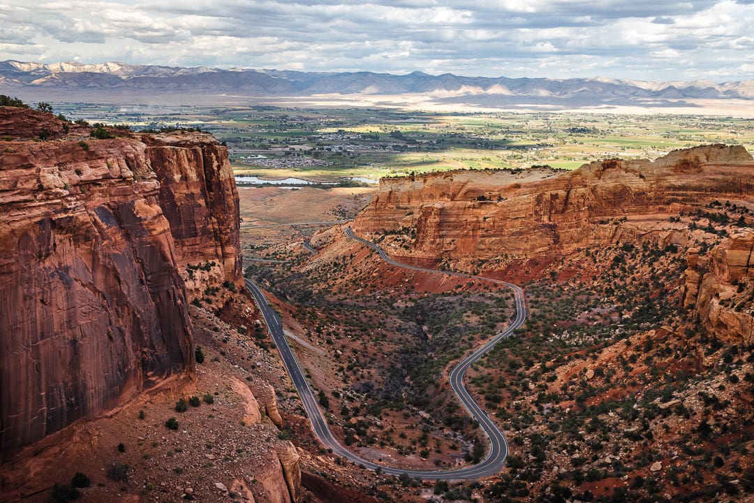 Winding road cuts through red rock canyon under a partly cloudy sky, with green fields and distant mountains in the background—a scene that perfectly captures the beauty of fall in Colorado.