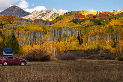 A red SUV with a rooftop tent is parked in a grassy field, surrounded by colorful autumn trees and distant mountains under a partly cloudy sky, capturing the vibrant beauty of Colorado fall.