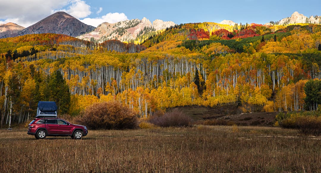 A red SUV with a rooftop tent is parked in a grassy field, surrounded by colorful autumn trees and distant mountains under a partly cloudy sky, capturing the vibrant beauty of Colorado fall.