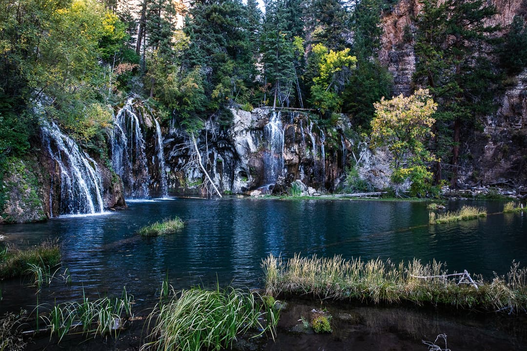 A serene pond with clear water, surrounded by lush green trees and rocky cliffs, with several small waterfalls cascading into the water from the forested hillside—this hidden oasis captures the magic of fall in Colorado, when vibrant autumn colors make it the best season to explore nature’s beauty.
