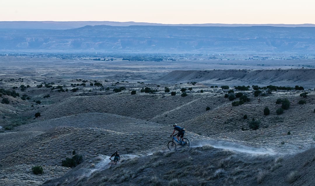 Two people ride mountain bikes on a dusty trail through rolling, dry hills at dusk, with expansive plains and distant mountains under a pale sky in the background—a perfect snapshot of fall in Colorado, when the landscape comes alive during the best season for outdoor adventure.