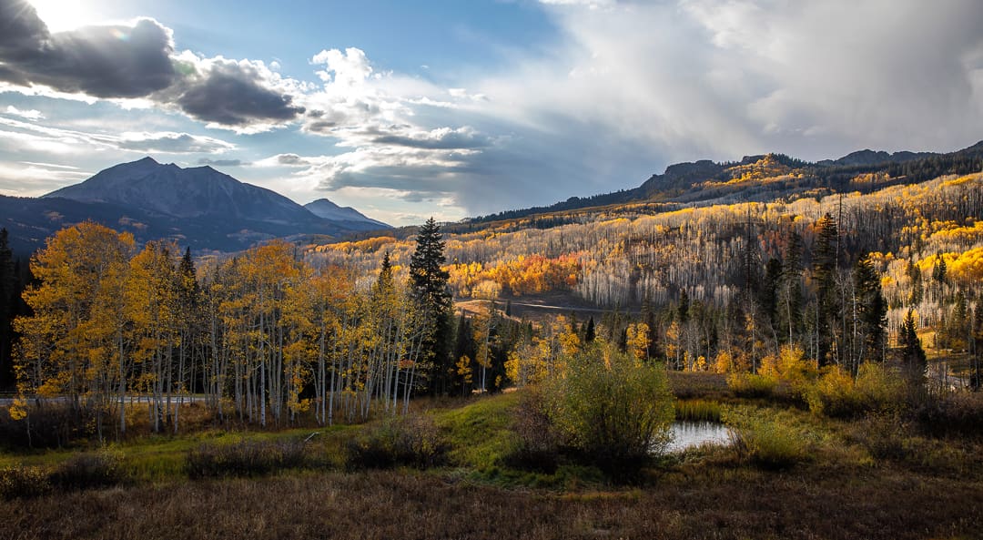 A scenic autumn landscape with mountains, yellow and orange trees, a small pond, and dramatic clouds lit by late afternoon sunlight captures the breathtaking beauty of fall in Colorado.
