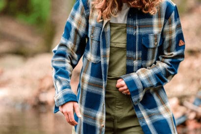 A person wearing a plaid shirt, olive waders, and a brown cap stands in a shallow creek, looking down. Their outfit resembles classic overalls, blending comfort and utility. Trees and rocks are blurred in the background.