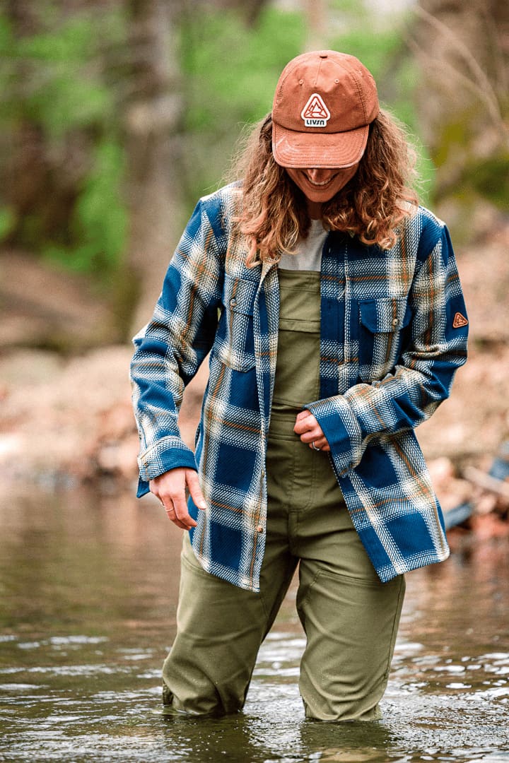 A person wearing a plaid shirt, olive waders, and a brown cap stands in a shallow creek, looking down. Their outfit resembles classic overalls, blending comfort and utility. Trees and rocks are blurred in the background.