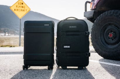 Two black suitcases, including an Aegis Carry-On, rest on the road next to a vehicle. A yellow sign in the background reads LOW FLYING AIRCRAFT. Mountains and a clear sky are visible behind the scene.