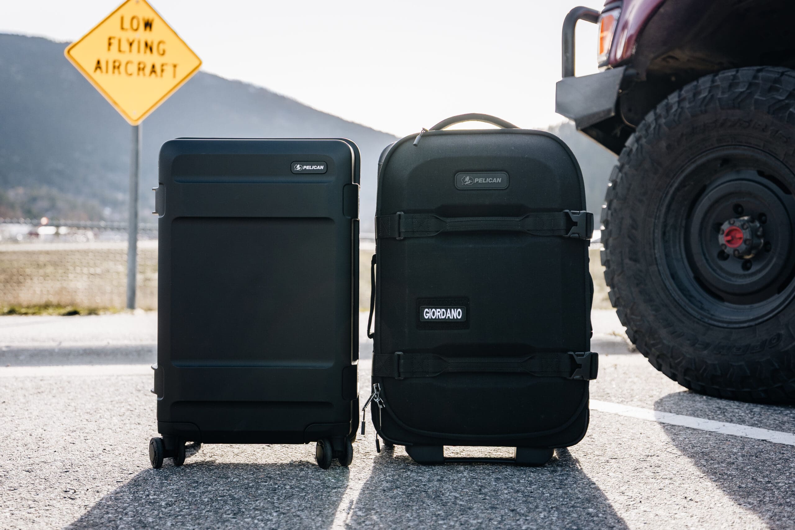 Two black suitcases, including an Aegis Carry-On, rest on the road next to a vehicle. A yellow sign in the background reads LOW FLYING AIRCRAFT. Mountains and a clear sky are visible behind the scene.