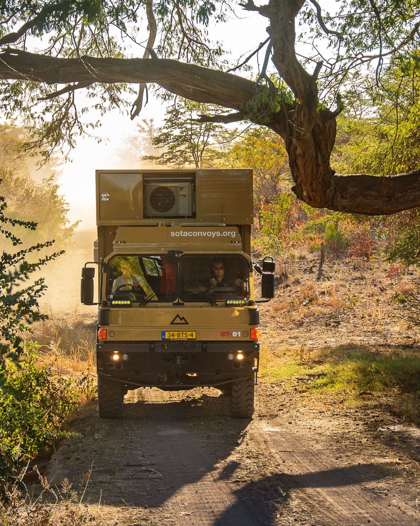 A tan expedition truck drives on a dirt path through a sunlit, wooded area, raising dust. Two people sit in the cab, perhaps searching for hidden desert oases. A large branch arches overhead.