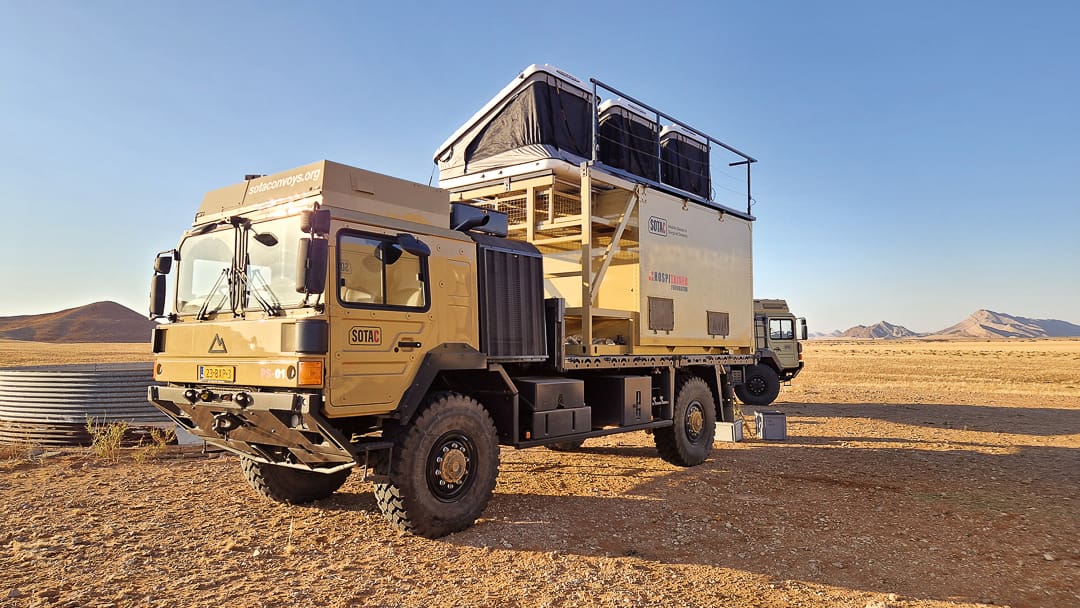 A large, tan expedition truck with an elevated camper box and open rooftop tent is parked in a desert landscape under a clear sky; mountains are visible in the background, evoking the spirit of mobile desert oases and OV conservation.