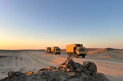 Three military trucks drive in convoy on a dirt road through a rocky desert landscape at sunset, with clear skies and scattered rocks in the foreground.