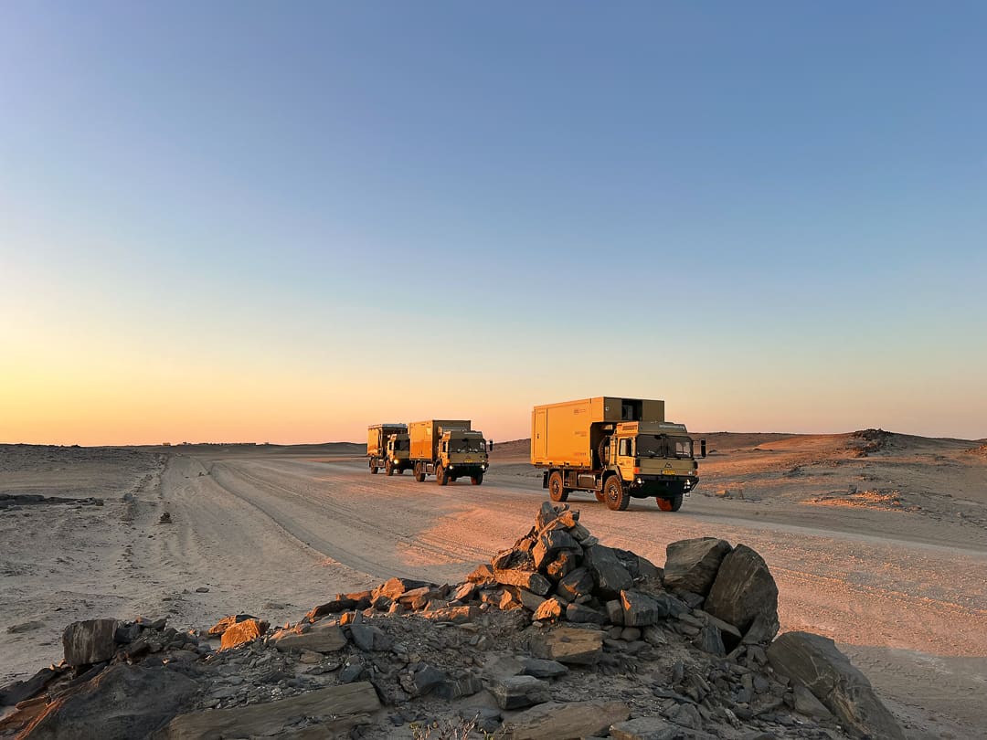 Three military trucks drive in convoy on a dirt road through a rocky desert landscape at sunset, with clear skies and scattered rocks in the foreground.