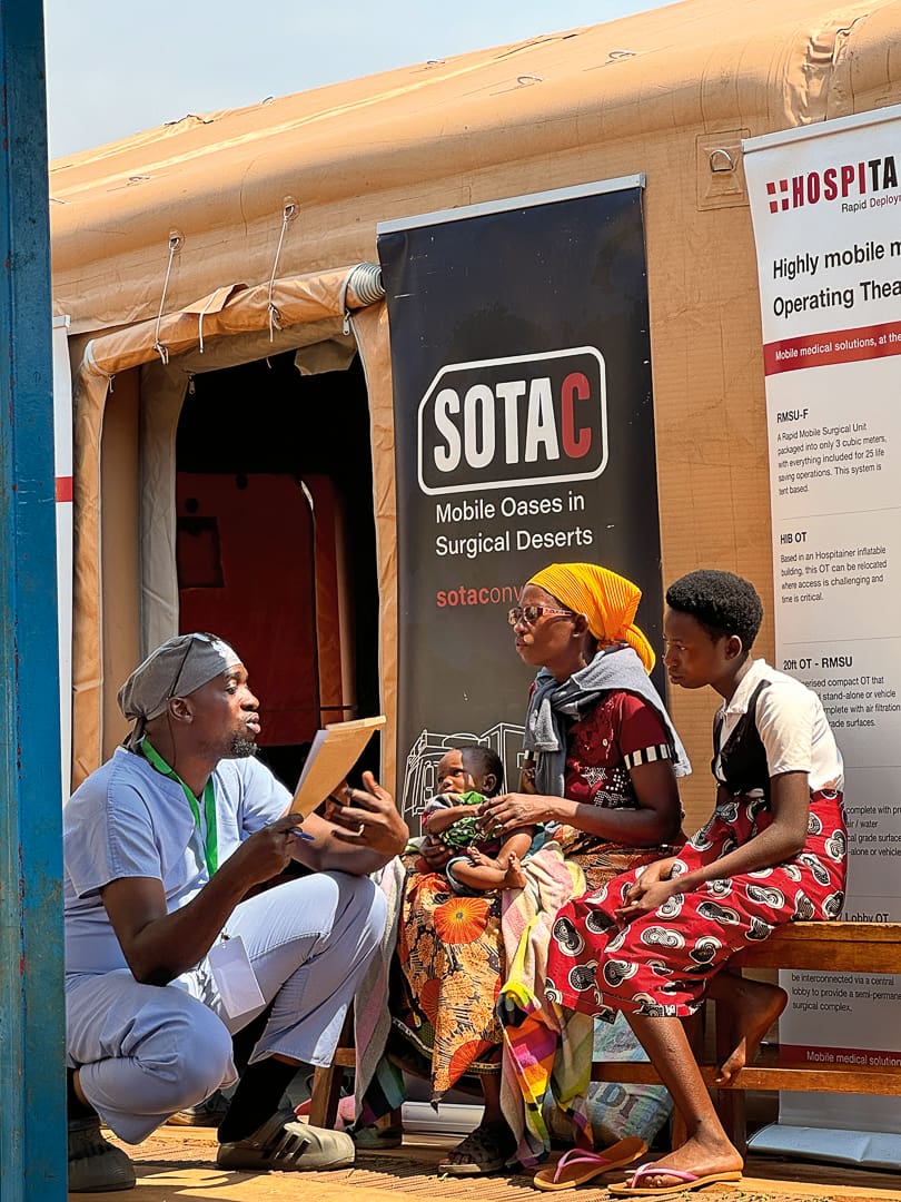 A healthcare worker talks to three people, including a child, outside a medical tent labeled SOTAC and Highly Mobile Operating Theatre. Set near an outdoor clinic, the group sits on benches as part of a medical convoy traveling between desert oases to provide essential care while supporting health conservation in remote communities.