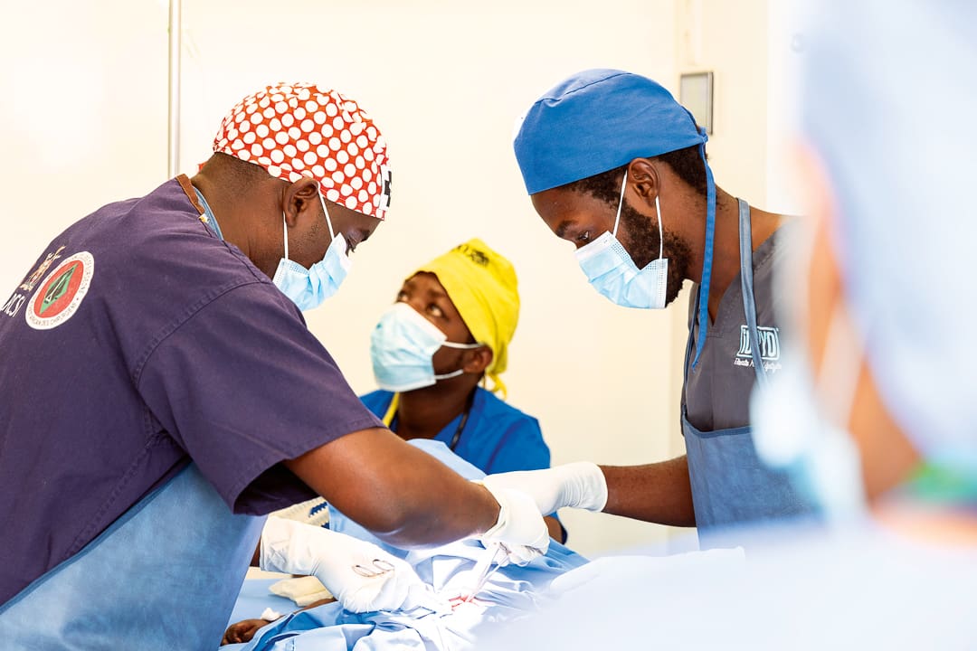 Three surgeons wearing scrubs, masks, and colorful caps perform an operation together in a brightly lit operating room, focusing intently on the procedure—working with the precision and coordination of medical convoys, dedicated to OV Conservation throughout every step.