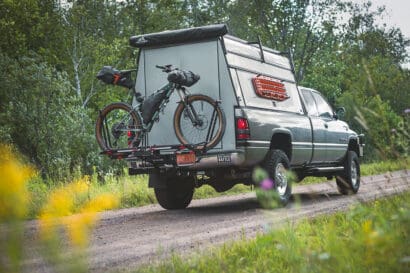 A pickup truck with a camper shell drives on a dirt road surrounded by greenery, embodying the spirit of escape. A bicycle is mounted on a rack at the back of the truck, ready for bikepacking adventures.