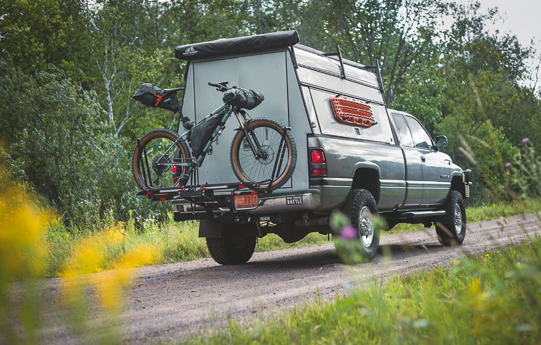 A pickup truck with a camper shell drives on a dirt road surrounded by greenery, embodying the spirit of escape. A bicycle is mounted on a rack at the back of the truck, ready for bikepacking adventures.