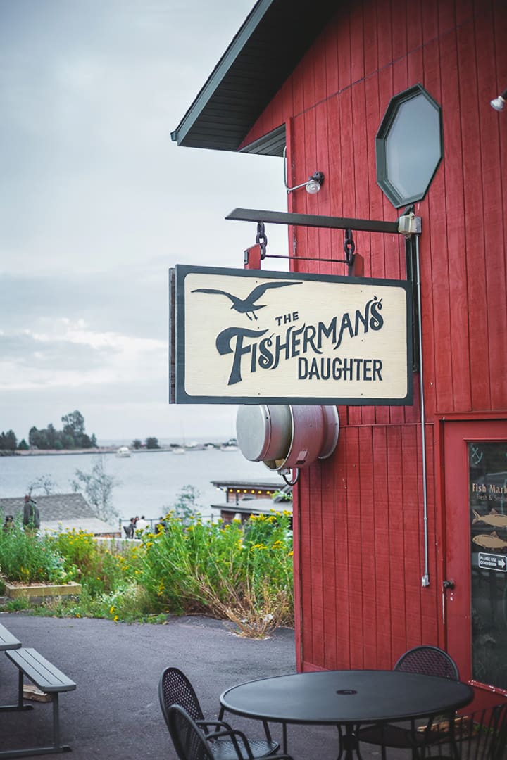 A wooden sign reading The Fishermans Daughter hangs on a red building near outdoor tables, with water and greenery visible in the background—an inviting stop for Minnesota bikepacking enthusiasts seeking adventure along scenic routes.