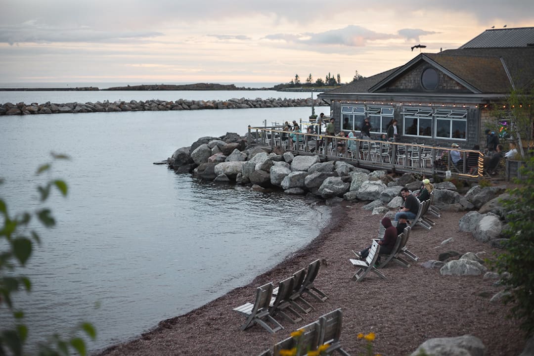 People relax on chairs along a rocky Minnesota lakeshore near a building with outdoor seating at sunset. String lights decorate the patio, and calm water stretches to the horizon—an inviting scene for anyone enjoying a bike adventure or stopping to unwind after a day of bikepacking through scenic trails.