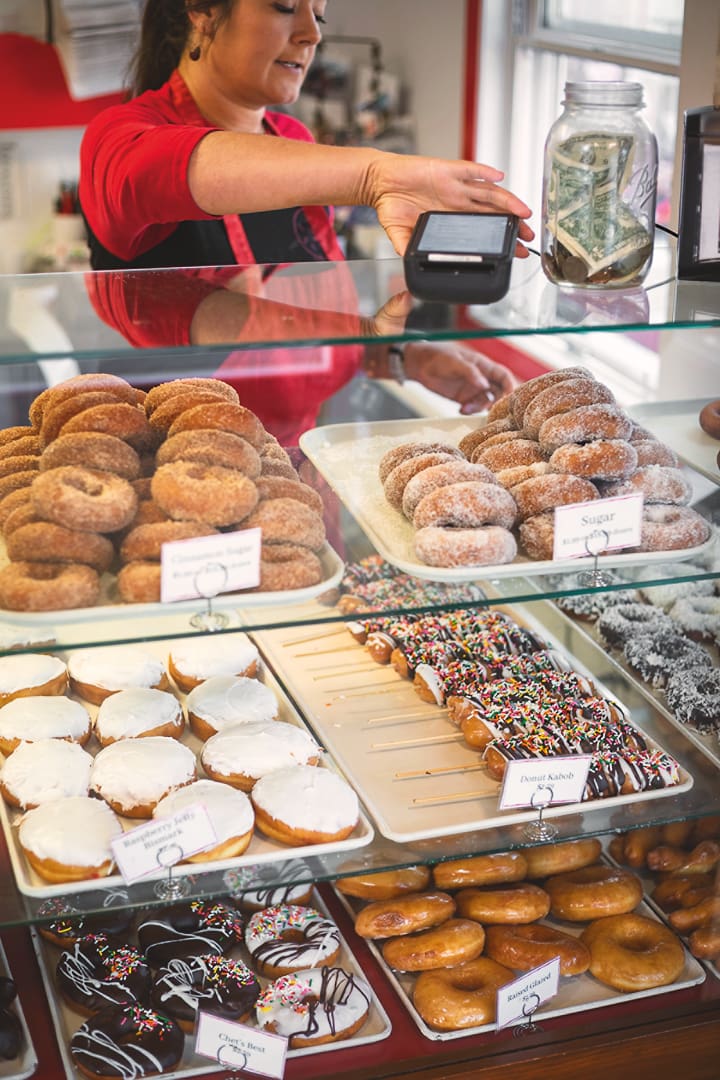 A person in a bakery rings up a purchase behind a glass display filled with donuts, cake pops, and pastries—a tempting escape for anyone exploring Minnesota or bikepacking through town. A tip jar with cash sits nearby.