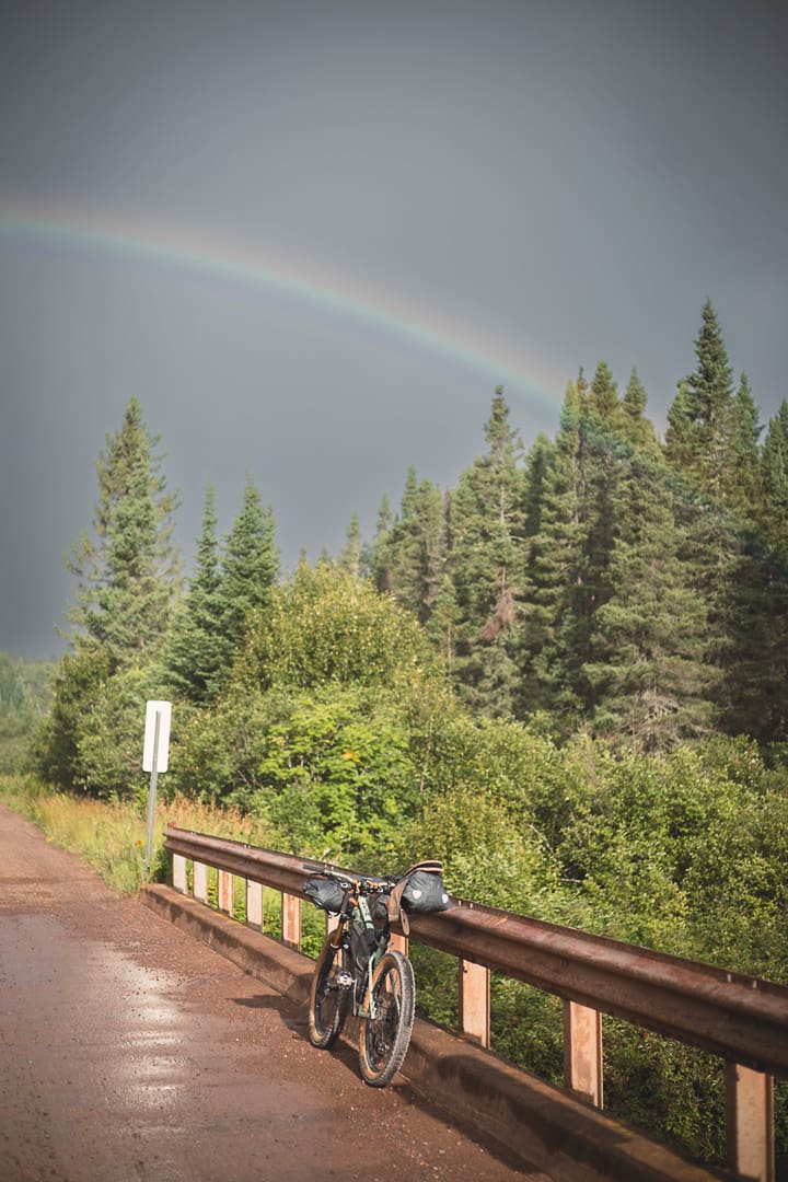 A bicycle leans against a guardrail on a wet road with tall pine trees in the background and a rainbow arching across a cloudy sky, capturing the spirit of bikepacking adventure in Minnesota.