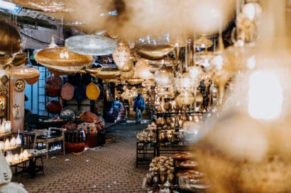 A marketplace filled with ornate hanging lamps and lanterns, with two people walking in the distance. Various colorful baskets, souvenirs, and wares line the cobblestone path under ambient lighting.