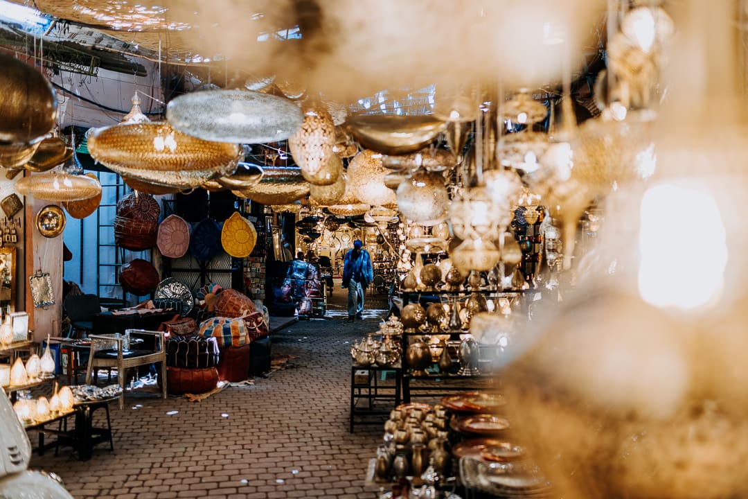 A marketplace filled with ornate hanging lamps and lanterns, with two people walking in the distance. Various colorful baskets, souvenirs, and wares line the cobblestone path under ambient lighting.