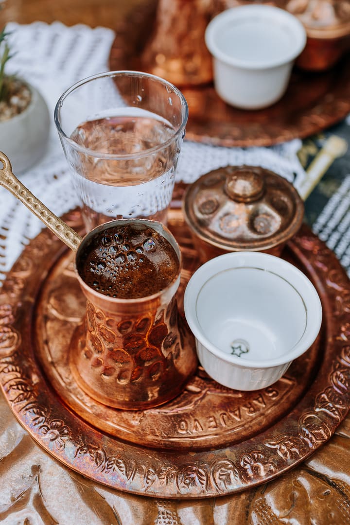 A traditional copper tray, often cherished as one of the finest souvenirs, holds a Turkish coffee pot, an empty porcelain cup, a glass of water, and a small lidded dish. The tray rests on a decorative tablecloth.