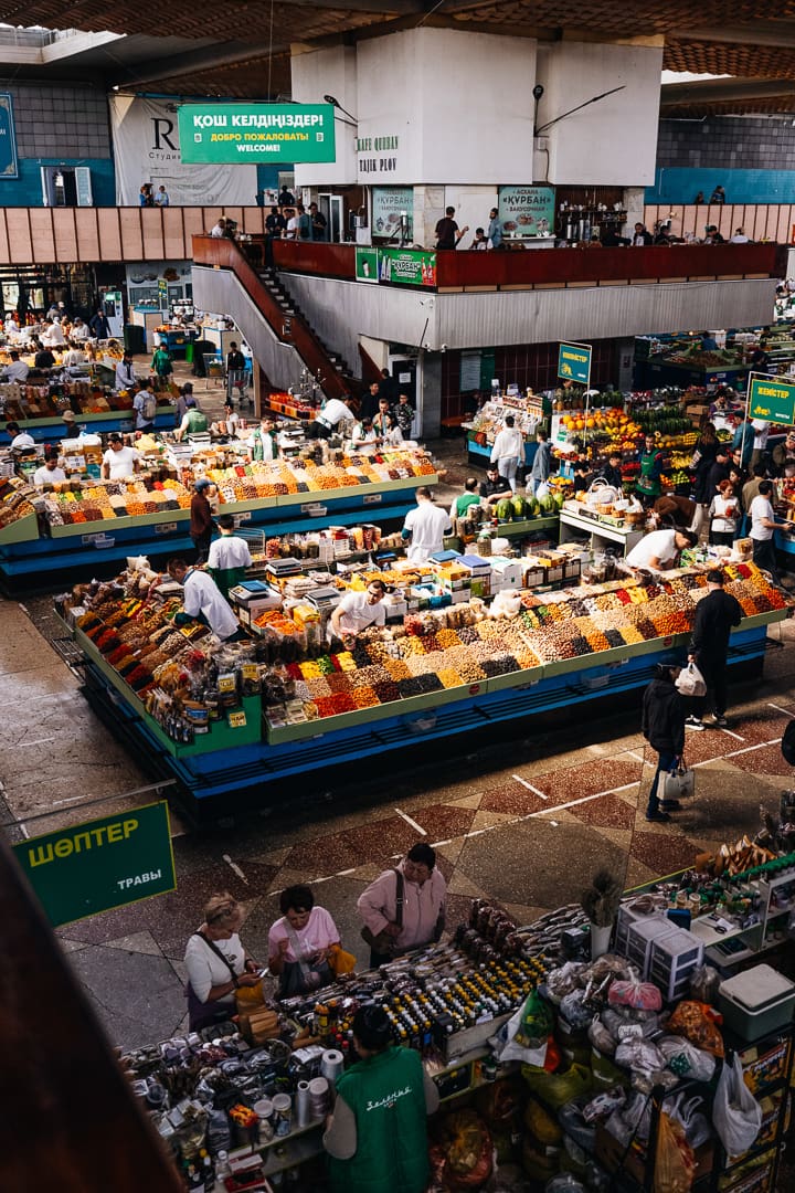 Indoor market scene with people shopping at colorful produce stalls, selling fruits, vegetables, souvenirs, and goods. Green signs with Cyrillic text hang above the busy, bustling area. Natural light filters in from windows above.