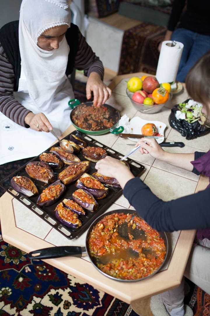 Two people prepare stuffed eggplants and a tomato-based filling at a table, perhaps exchanging stories about their favorite souvenirs from past travels. Fresh fruits, salad, and paper towels are nearby. One person wears a white headscarf, while the other reaches for the eggplants.