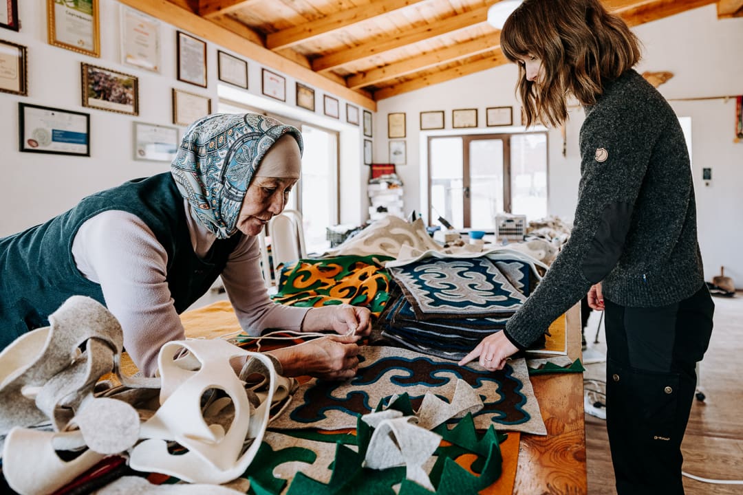 Two women work on colorful felt fabric crafts, creating unique souvenirs at a wooden table in a room decorated with certificates on the wall, natural light pouring in through large windows.