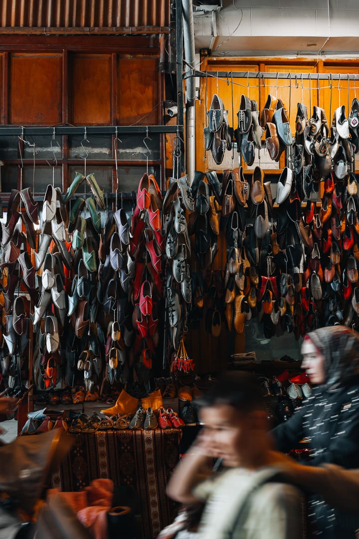 A market stall displays many pairs of shoes, sandals, and colorful souvenirs hanging, with a blurred person passing by in the foreground and another person in a headscarf on the right. The scene is busy and vibrant.