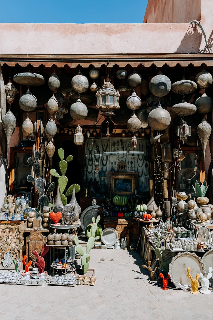 Outdoor market stall displaying metal lanterns, trays, plates, and decorative items, with colorful cacti ornaments and unique souvenirs alongside other crafts arranged on tables and shelves in front of a rustic building under a clear blue sky.