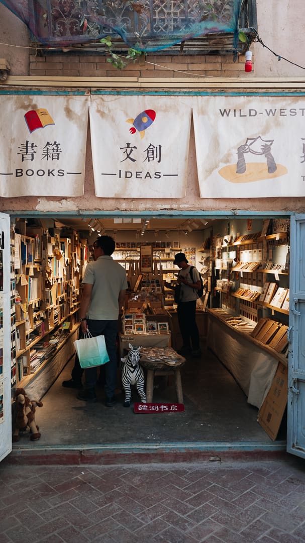 A small bookstore with open doors shows two people browsing books inside. The store has banners with Chinese and English words for Books and Ideas. A toy zebra is on a table at the entrance, surrounded by an assortment of souvenirs that invite visitors to take home a memory from their visit.
