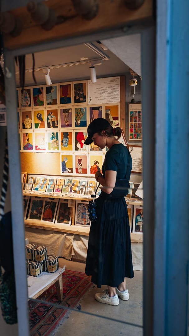 A woman in a black dress and cap looks at art prints in a cozy gallery, surrounded by colorful illustrations and unique souvenirs displayed on the walls and shelves.