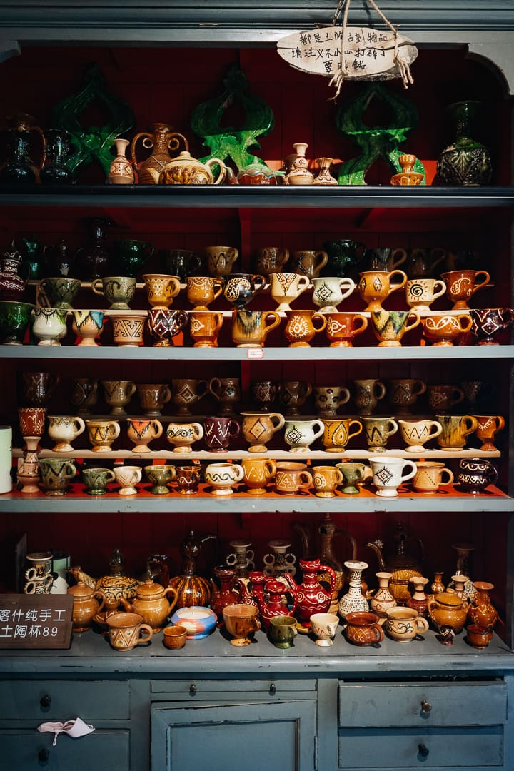 A wooden shelving unit filled with rows of colorful handmade ceramic mugs, jugs, and pots with various patterns and designs, arranged neatly against a dark-red background—each piece offering a charming selection of unique souvenirs.