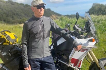 A man wearing sunglasses and a cap stands outdoors next to a touring motorcycle with luggage, surrounded by greenery and hills under a partly cloudy sky—a fitting tribute for Arden Kysely (1953-2026), captured in this moment of quiet reflection during his Celebration of Life.