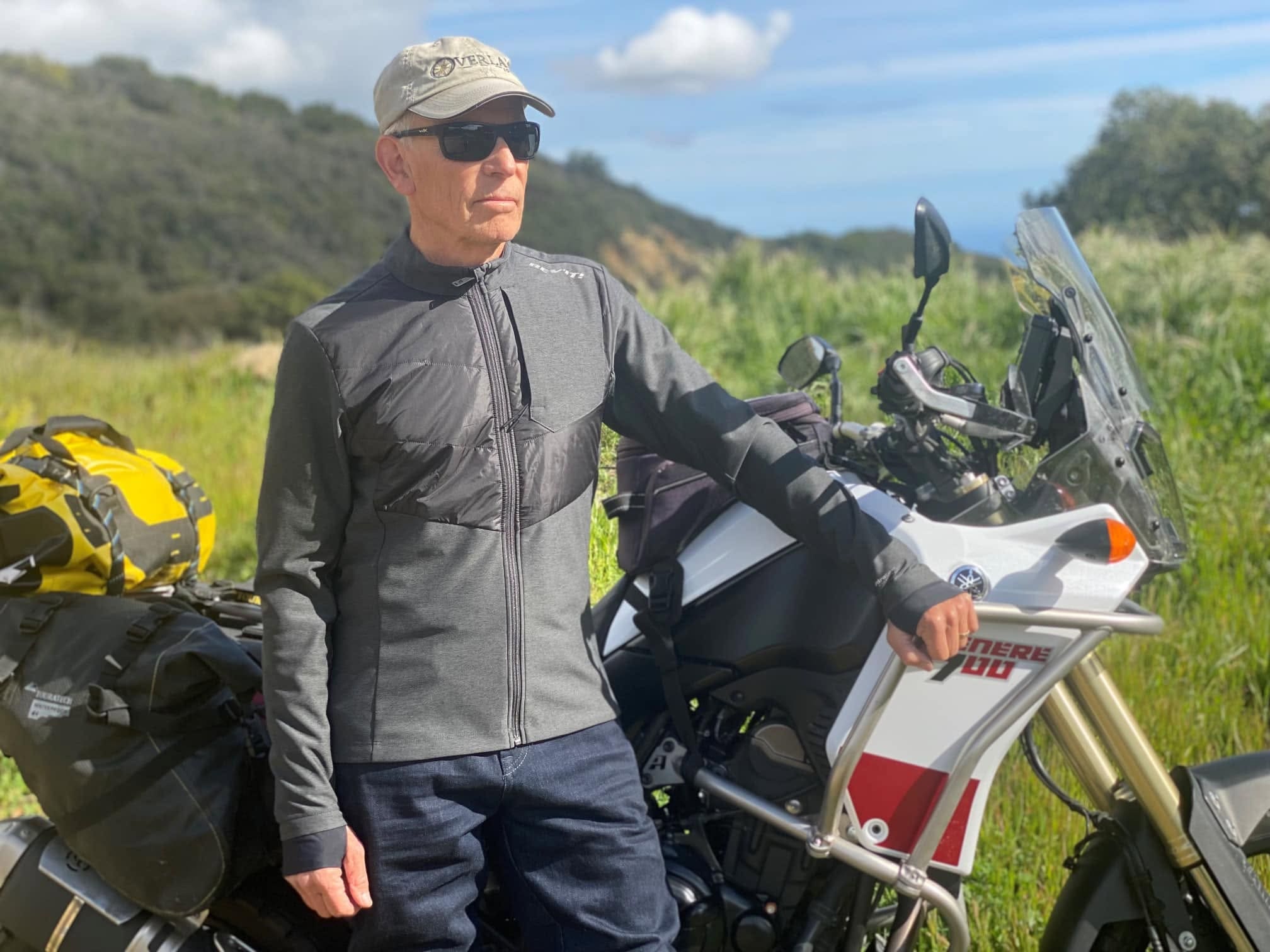A man wearing sunglasses and a cap stands outdoors next to a touring motorcycle with luggage, surrounded by greenery and hills under a partly cloudy sky—a fitting tribute for Arden Kysely (1953-2026), captured in this moment of quiet reflection during his Celebration of Life.