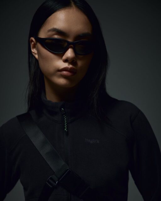 A person with long dark hair wearing black sunglasses, a black jacket, and a crossbody strap looks slightly to the side against a dark background, embodying the bold style often featured in Overland News.