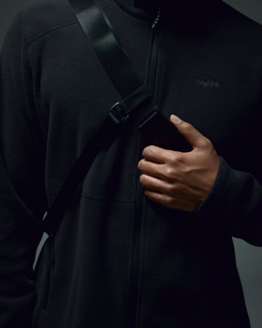 A person wearing a black zip-up jacket holds a black strap across their chest with their right hand. The image, reminiscent of an Overland News feature, focuses on their torso and hand, not showing their face.