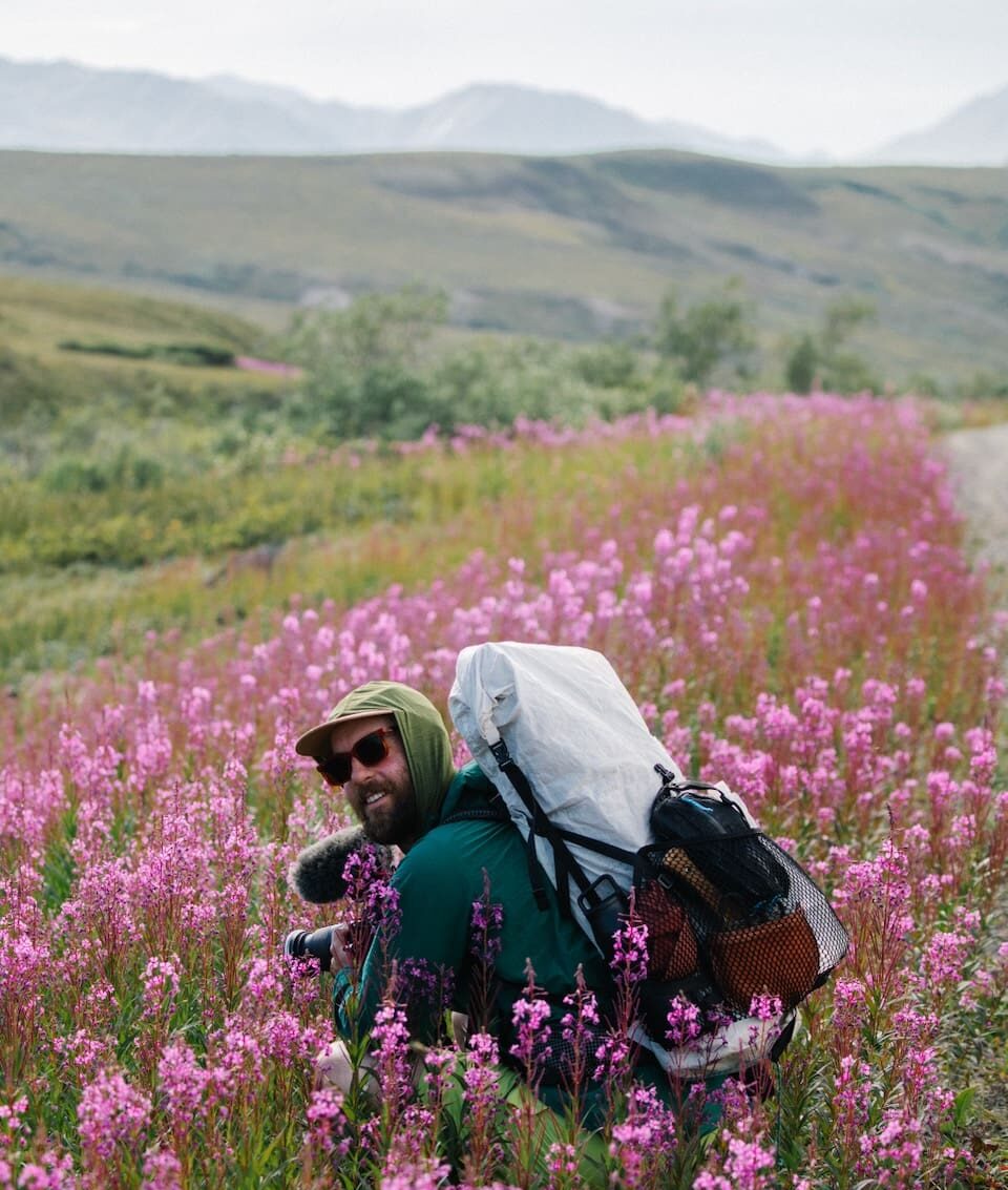 A person wearing a green jacket and backpack sits in a field of blooming pink wildflowers, smiling and looking at the camera with mountains and greenery in the background.