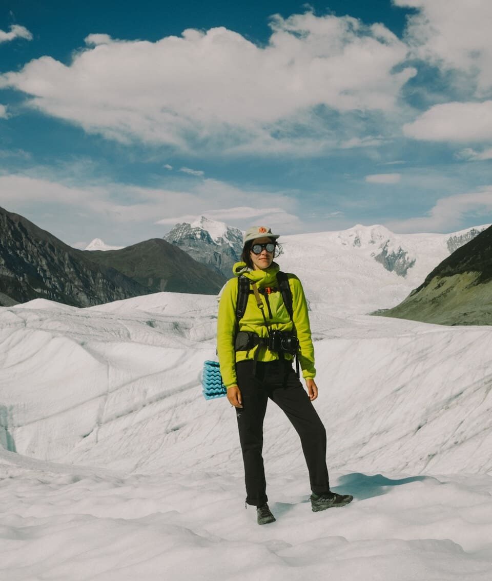 A person in hiking gear stands on a snowy glacier with mountains and blue sky in the background.