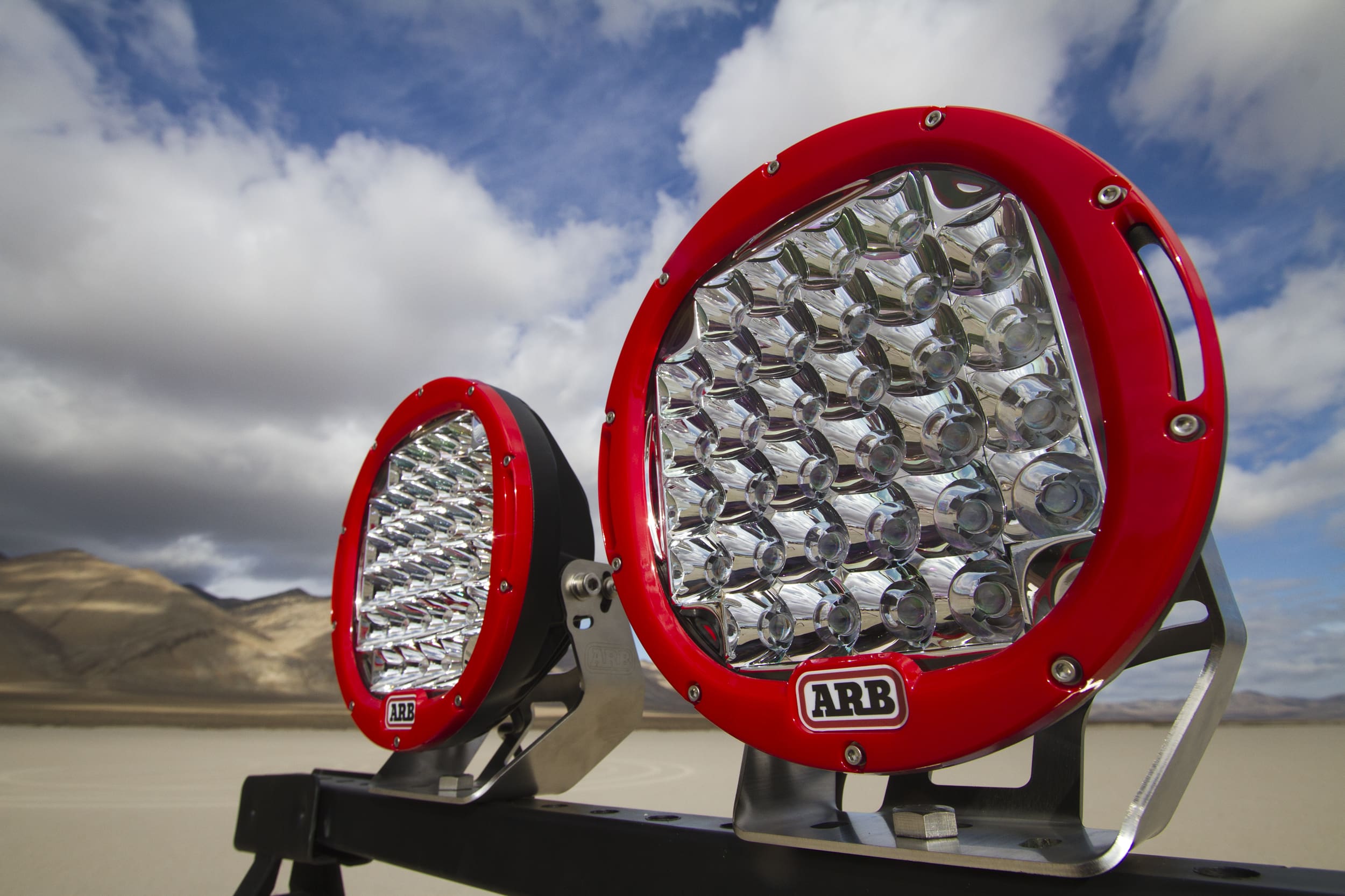 Two round, red-rimmed ARB LED driving lights are mounted on a vehicle against a backdrop of cloudy blue sky and sandy desert landscape.