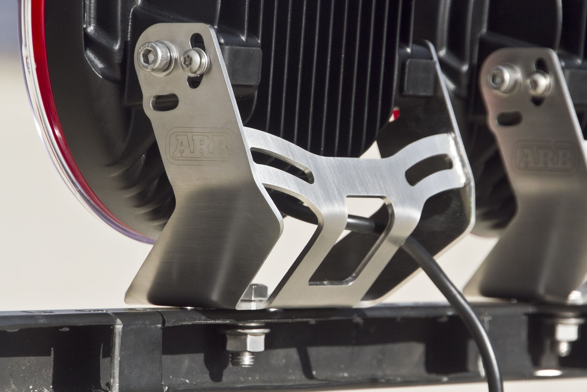 Close-up of a metal ARB light mounting bracket attached to a vehicle, supporting robust driving lights. A black cable runs from the mounted device, and the bolts and detailed metalwork are visible.