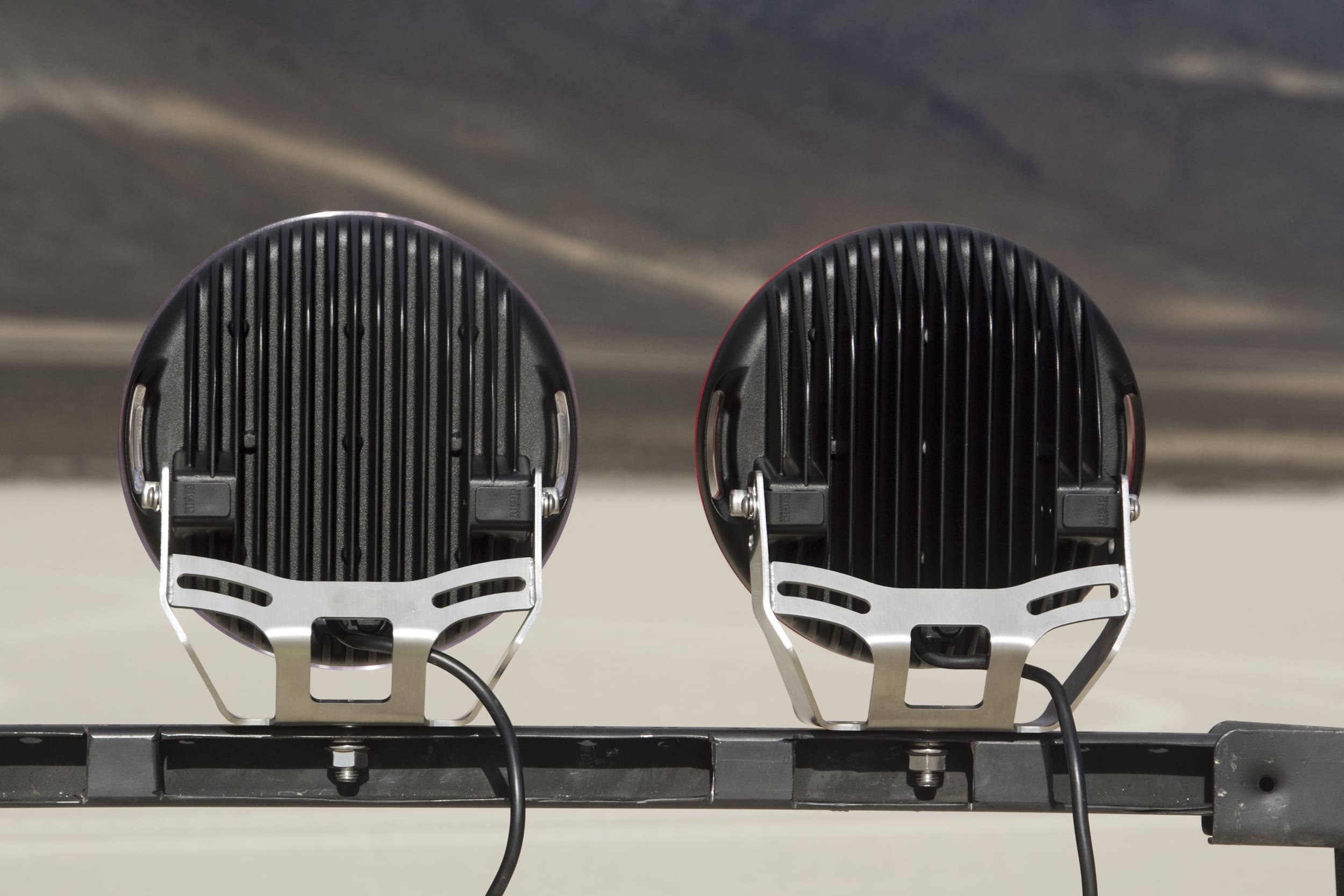 Two round black driving lights are mounted side by side on a metal bar, shown from the back. The image background is out of focus, displaying a flat, pale surface and dark hills.