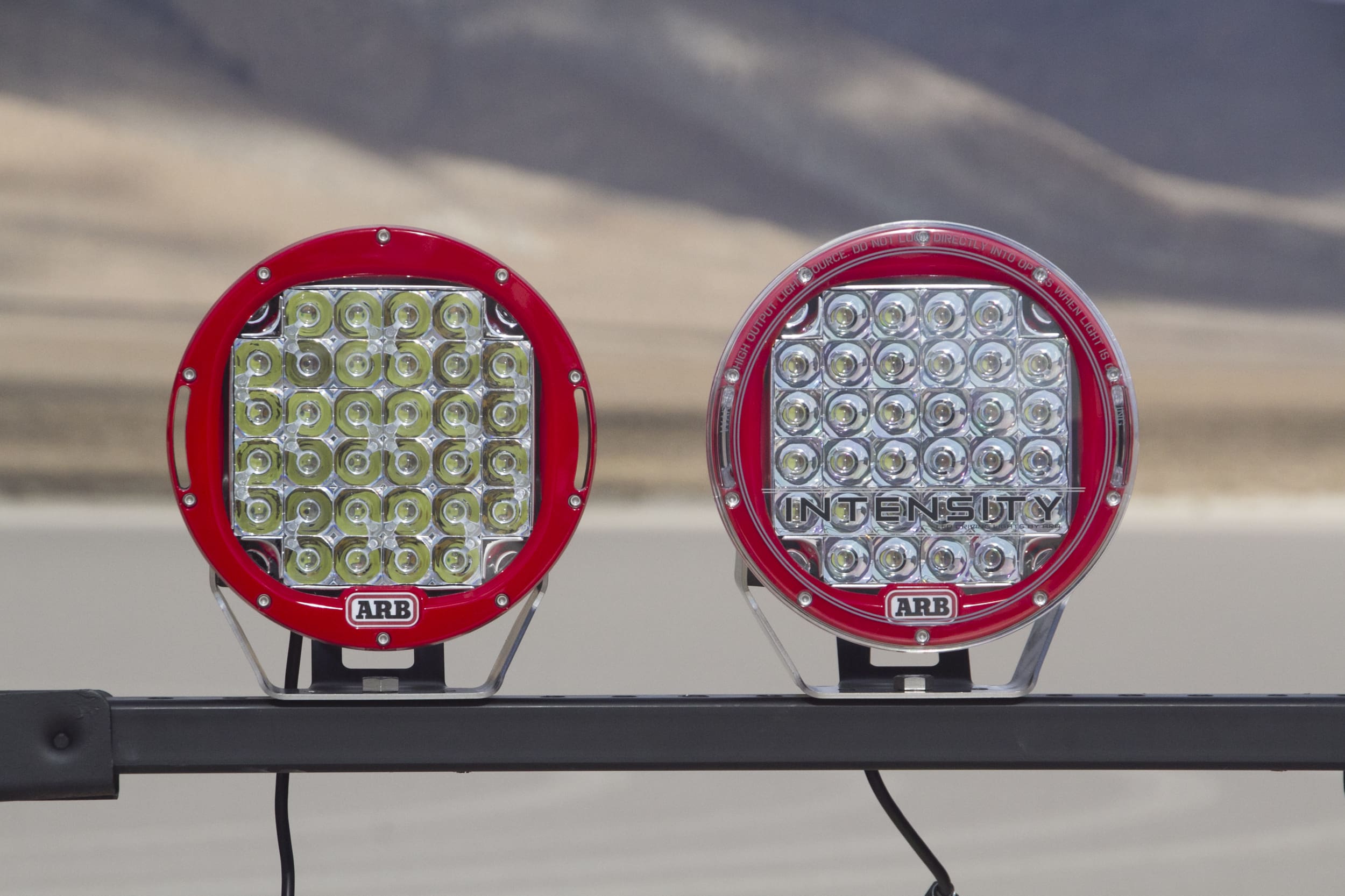 Two round red ARB off-road driving lights are mounted side by side on a metal bar, set outdoors with a blurred, arid landscape and mountains in the background.
