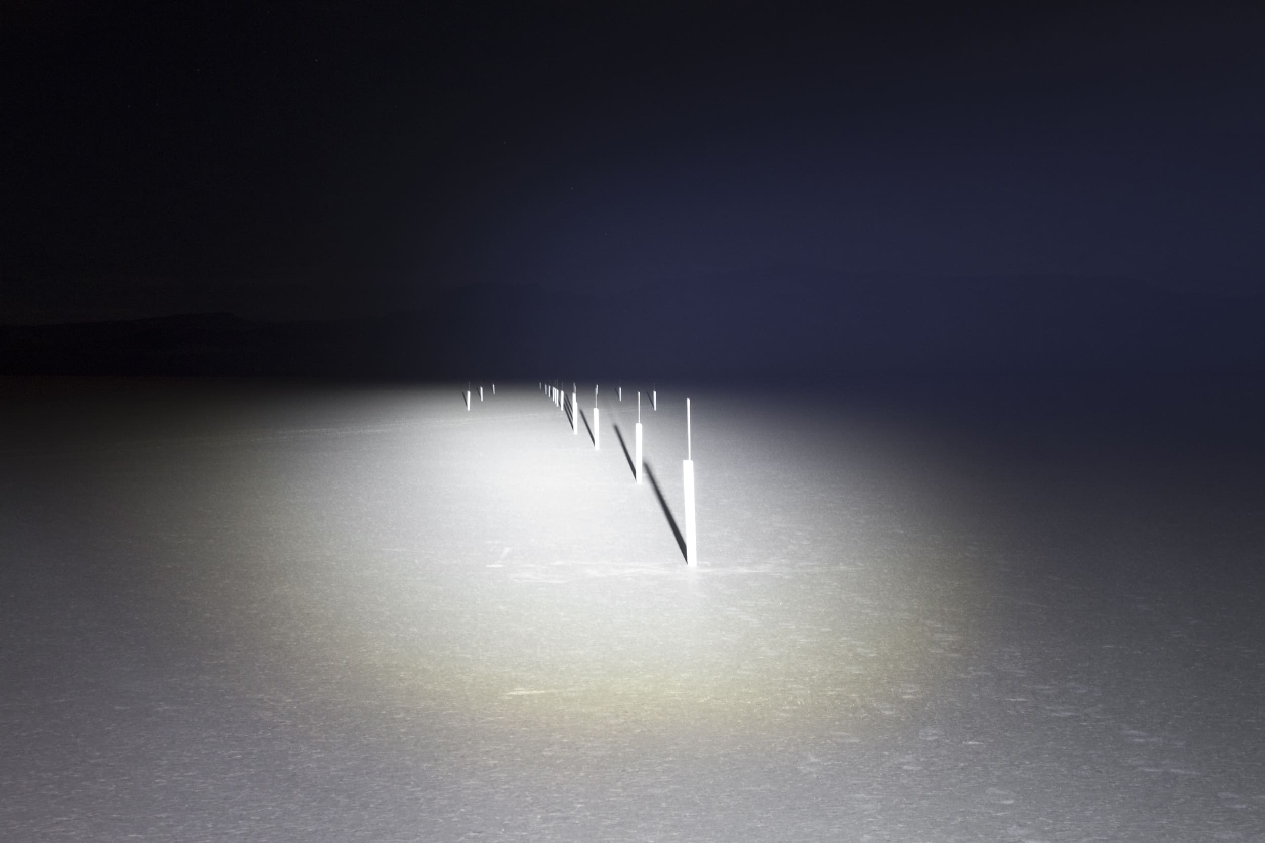 A row of white poles illuminated by driving lights stretches into the distance on a flat, dark landscape at night.