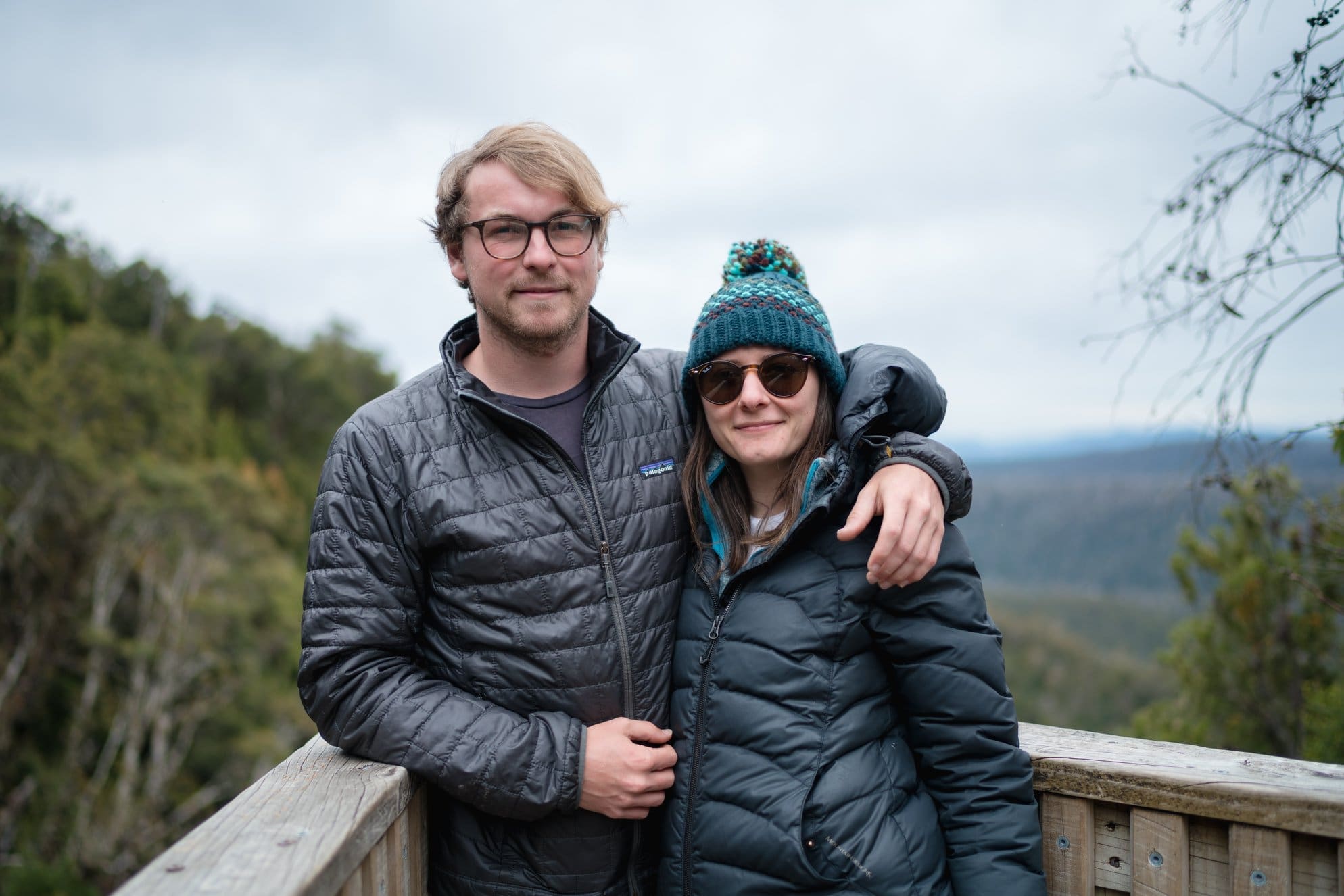 A man and woman in winter jackets and sunglasses stand on a wooden deck with scenic forest and hills in the background. The man has his arm around the woman, and both are smiling at the camera.